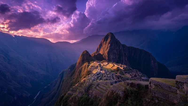 Dramatic aerial view of Machu Picchu during golden hour sunset with the ancient citadel illuminated against purple mountain silhouettes and clouds