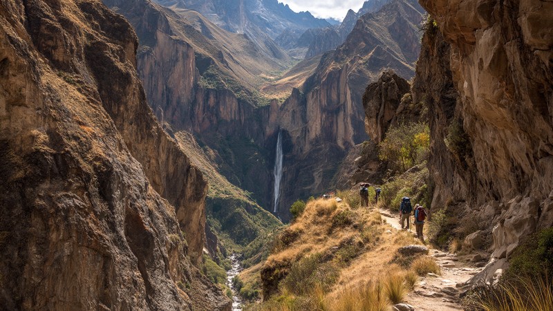 Hikers walking along a narrow trail in Peru’s Cotahuasi Canyon with towering cliffs and a waterfall in the distance