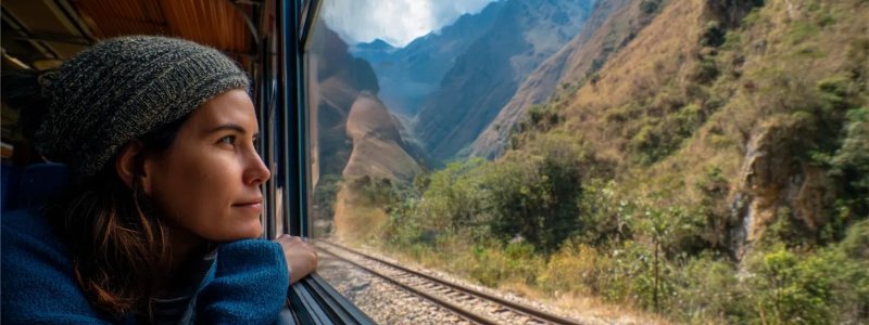 Professional traveler sitting on train from Cusco to Aguas Calientes looking out window at Andean mountain scenery with railroad tracks visible