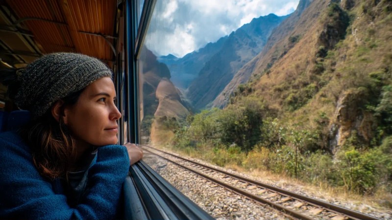 Professional traveler sitting on train from Cusco to Aguas Calientes looking out window at Andean mountain scenery with railroad tracks visible