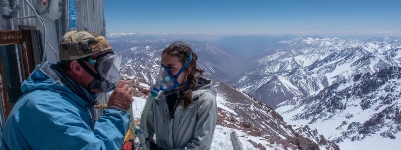 Traveler experiencing symptoms of altitude sickness with oxygen mask at high altitude medical station with snow-capped Andes mountains in background