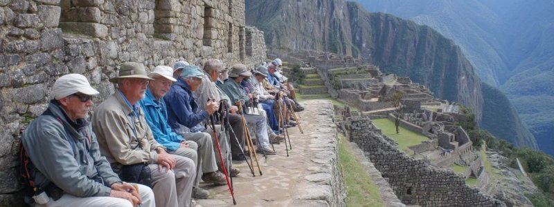Senior travelers aged 60+ enjoying Machu Picchu with comfortable transportation, walking at a relaxed pace, using walking sticks, sitting on benches while admiring views, and participating in age-appropriate activities with professional guide assistance
