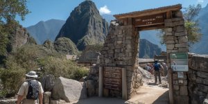 Tourist looking at Machu Picchu entrance sign showing operating hours and visitor capacity numbers with ancient citadel visible in mountain background