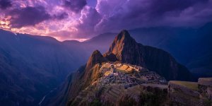 Dramatic aerial view of Machu Picchu during golden hour sunset with the ancient citadel illuminated against purple mountain silhouettes and clouds