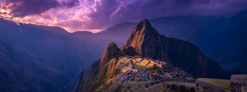 Dramatic aerial view of Machu Picchu during golden hour sunset with the ancient citadel illuminated against purple mountain silhouettes and clouds