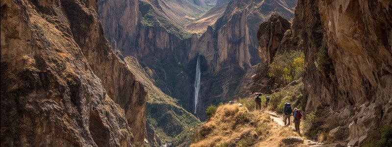 Hikers walking along a narrow trail in Peru’s Cotahuasi Canyon with towering cliffs and a waterfall in the distance