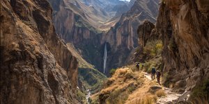 Hikers walking along a narrow trail in Peru’s Cotahuasi Canyon with towering cliffs and a waterfall in the distance